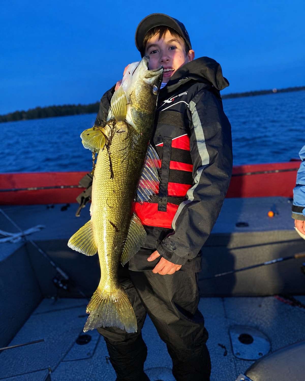 A child in a cap, rain jacket and life jacket holding up a large fish in a boat under a deep blue twilight sky