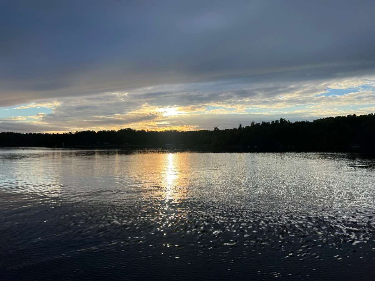 The sun breaking through clouds above the wooded far shore, its light reflected in a long path across the rippled lake